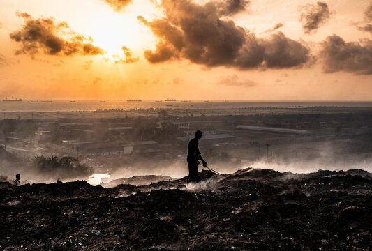 Chattogram, Bangladesh - 17 October 2026: View of a worker sorting through waste at a landfill site under a dramatic sunset sky with cargo ships visible on the horizon.