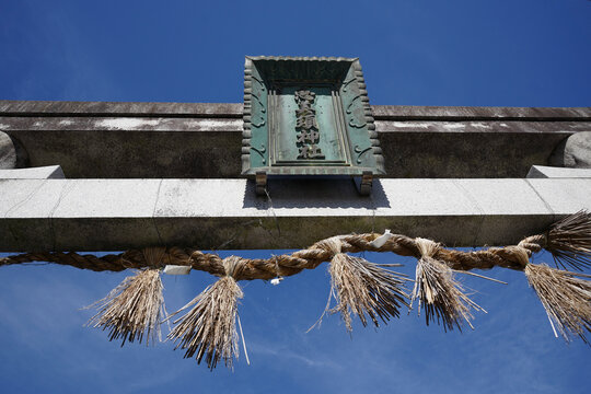 Low Angle View of Stone Torii Gate with Bronze Shrine Plaque and Shimenawa Sacred Straw Rope Against Blue Sky