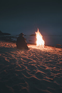 Merikula, Harju maakond, Estonia - 29 August 2020: View of a person sitting by a bonfire on the sandy beach at night with the glowing flames illuminating the shore.