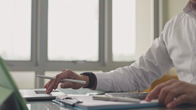 Businessman uses touchpad on laptop while holding stylus near paperwork in corporate office. Financial analytics audit, project workflow management, digital executive strategy documentation.