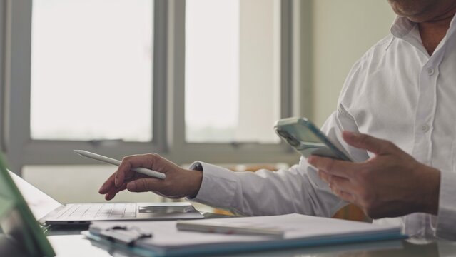 Businessman checks mobile phone while typing on laptop computer at office desk. Corporate communication workflow, financial report analysis, digital marketing management.