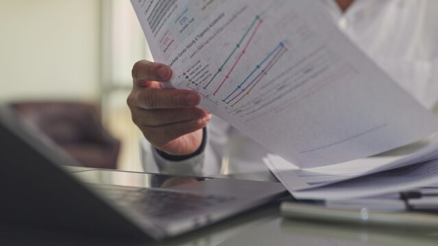 Businessman analyzes statistical chart paperwork beside laptop on office desk. Financial performance audit, market research data analysis, corporate investment forecasting strategy review.