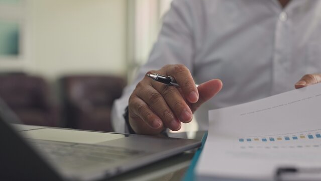 Businessman reviews financial chart report beside laptop computer in office workplace. Corporate audit paperwork analysis, investment revenue accounting review, annual sales budget verification.