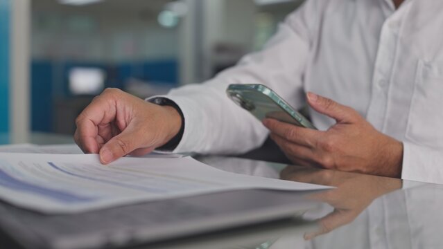 Businessman reviews paper financial report documents while holding mobile phone at office desk. Corporate accounting audit process, mobile banking data verification, investment paperwork analysis.