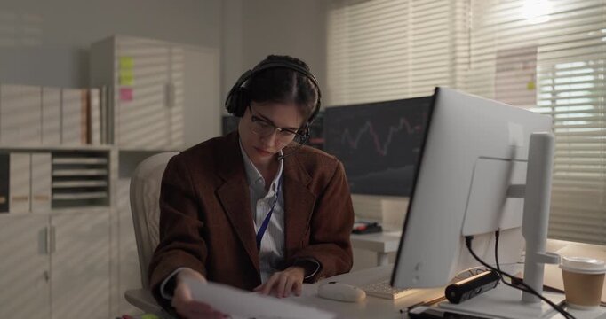 Woman stock trader in headset reading printed report at desk with market charts on monitor