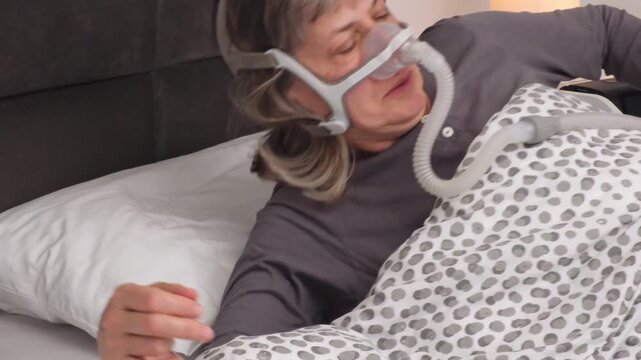 Middle-Aged Woman with Graying Hair Waking Up in Bed and Removing CPAP Mask After Using Sleep Apnea Device During the Night.
