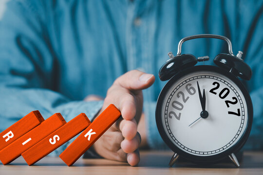 Hand stopping falling wooden domino blocks beside an alarm clock showing 2026 to 2027 symbolizing business planning and crisis protection of risk management.