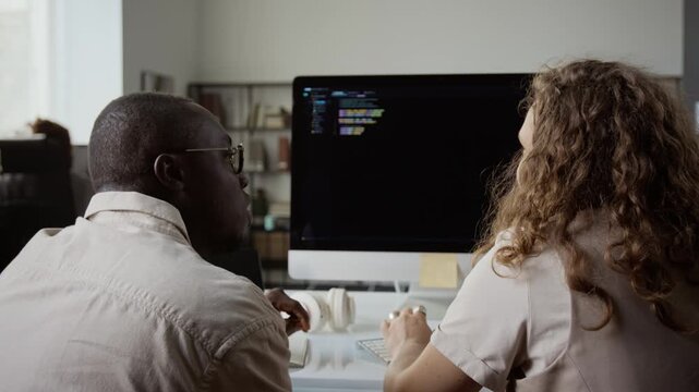 African American young adult man and Caucasian young adult woman coding on desktop computer from rear view. Footage ideal for software engineering, IT recruiting and tech advertising