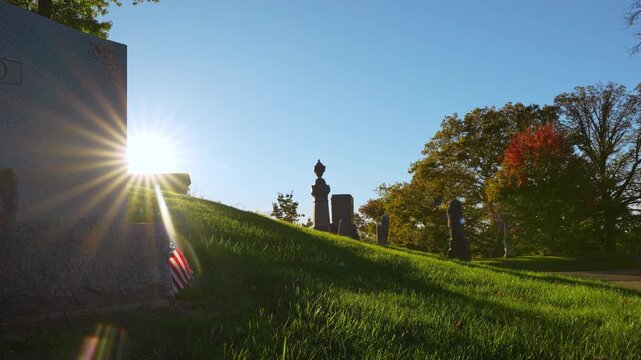 Sunlight forms a starburst beside a grave monument on a grassy hill. An American flag rests near the shaded stone as autumn trees line the right side beneath a blue sky.