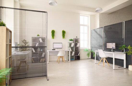 Modern office workspace with desks and computers. Bright interior with dividers, minimalist shelving and plants, flooded with daylight from tall windows. Useful for corporate web and print.