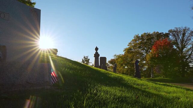 The sun shines past a headstone above a cemetery slope. Radiating beams cross the left foreground while green grass and orange leaves fill the hillside.