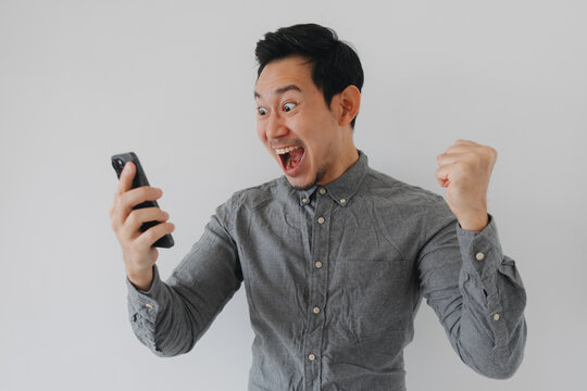 Excited Asian man celebrating success with a fist pump while looking at his smartphone.