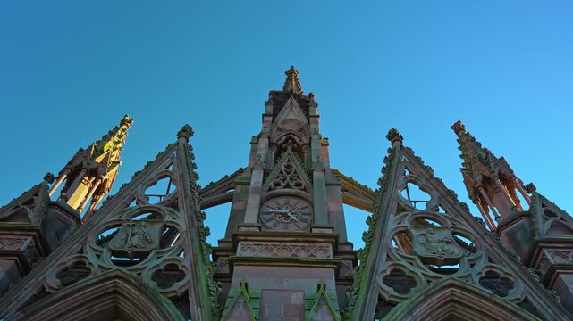 Gothic pinnacles rise above the cemetery entrance in Brooklyn. Carved tracery and pointed gables appear against a blue sky from below.