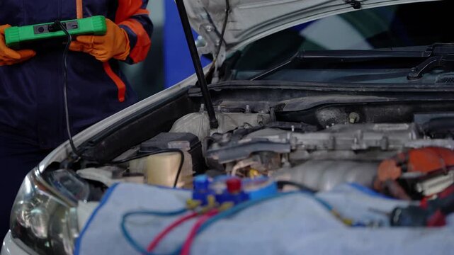 Engine bay of a white car with the hood open and blue fender cover, while a technician in orange gloves walks in holding a green diagnostic scanner on a long cable