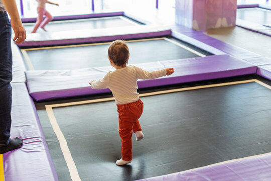 Toddler Bouncing on Trampoline with Arms Out While Father Watches, Back View