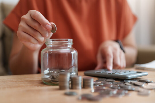 Woman dropping a coin into a glass savings jar beside a calculator and stacks of coins on a wooden table. Represents personal savings, budgeting and money management.