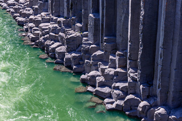 Green river currents flow beside tall basalt columns in Studlagil canyon, East Iceland. Sunlight picks out angular rock ledges and ripples along the shadowed cliff base. © Artem