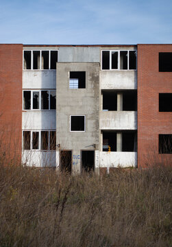 Unfinished concrete and brick building with empty window openings amid overgrown grass. Incomplete construction site with exposed structure. Urban development and building project.