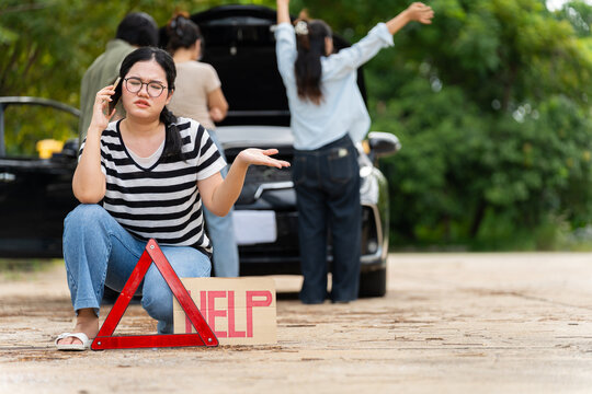 An Asian woman speaks on a phone beside a roadside warning triangle as companions inspect their car, showing stress from a vehicle breakdown and the need for emergency or insurance assistance.