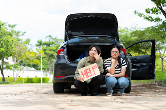 Two Asian women sit beneath an open car trunk holding a help sign on a tree-lined roadside, illustrating a breakdown, stranded travelers, and the need for roadside assistance and insurance.