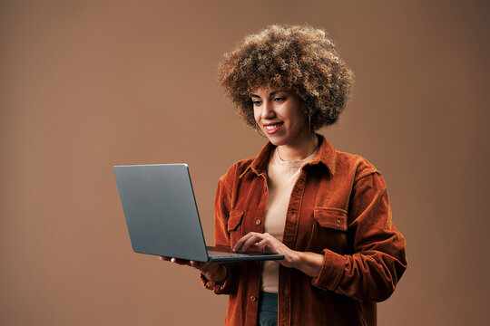 Smiling young woman with curly hair using a laptop against a warm brown studio background, showing remote work, online learning and modern technology.