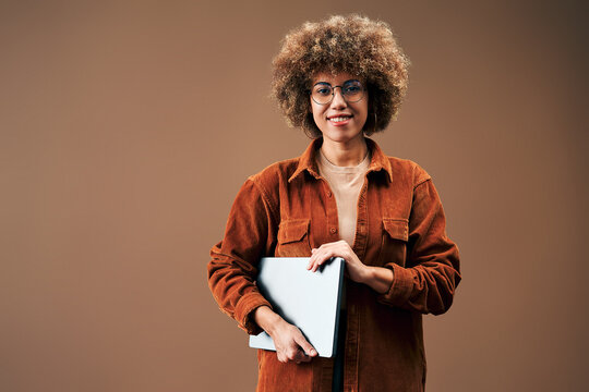 Smiling woman with curly hair and glasses holding a laptop against a warm brown studio background, conveying confidence, creativity, and modern professional style.