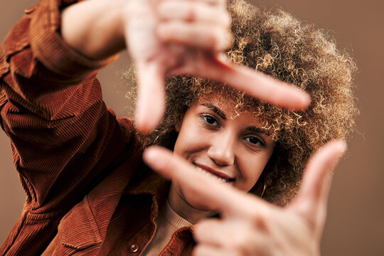 Smiling young woman with curly hair frames her face with her hands against a warm brown background, creating a playful creative portrait