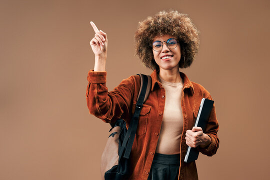 Smiling young woman with curly hair and glasses points upward while holding a notebook and wearing a backpack against a warm brown studio background.