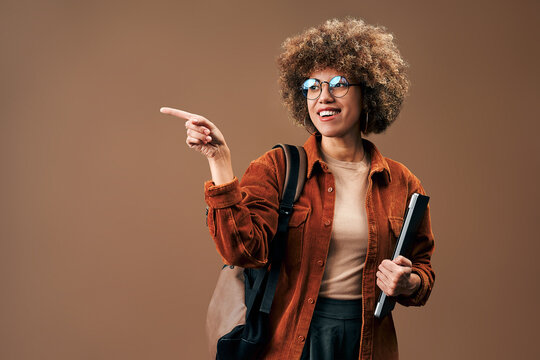 Smiling young woman with curly hair, glasses and backpack points aside while holding a notebook against a warm brown studio background.