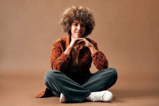 Smiling young woman with curly hair sits cross-legged on the floor in casual clothes against a warm brown studio background.