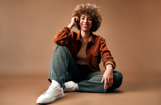 Smiling young woman with curly hair sits casually on a studio floor in warm brown tones, wearing an orange jacket, jeans, and white sneakers.