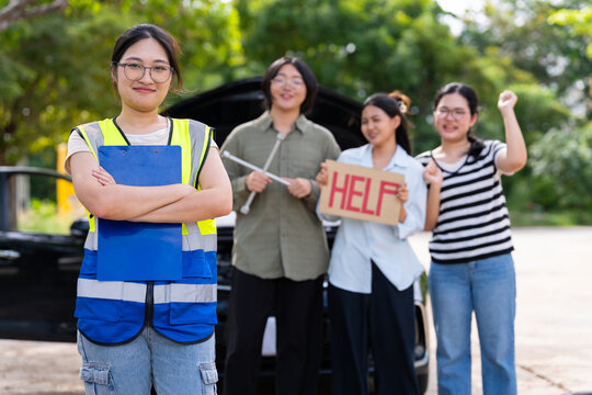 A female roadside worker poses with a clipboard while three women stand beside a car with its hood open, presenting vehicle breakdown assistance, teamwork, and women in automotive services.