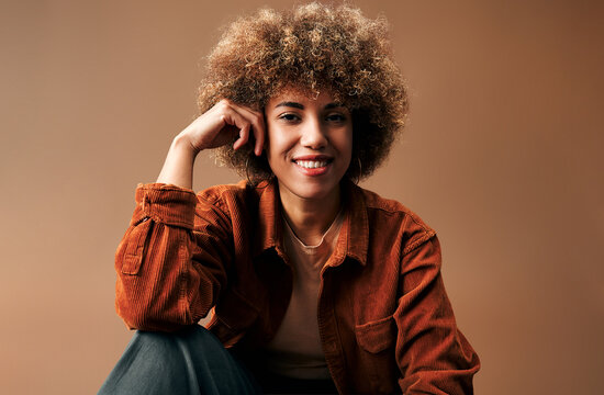 Joyful young mixed-race woman with curly hair smiling against a warm brown background.