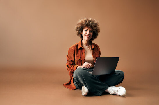 Smiling young woman with curly hair sits cross-legged on a warm brown floor while using a laptop, creating a relaxed scene for work, study, or online communication.
