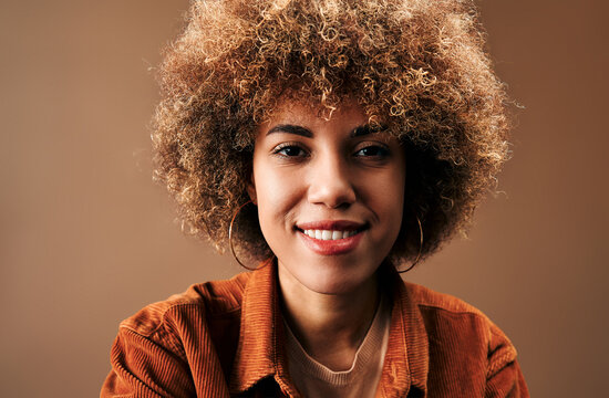 Close-up studio portrait of a smiling young woman with natural curly afro hair, warm brown skin, hoop earrings and an orange corduroy jacket against a soft brown background.