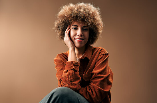Warm studio portrait of a smiling young woman with curly hair wearing a rust-colored corduroy shirt, resting her face on her hand against a brown background.