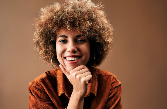 Smiling young woman with natural curly hair resting her chin on her hand, wearing a rust corduroy jacket against a warm brown studio background.