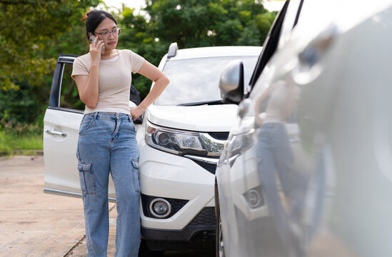Asian woman speaks on a mobile phone beside a white car while another vehicle fills the foreground, suggesting a possible collision and the need to contact insurance or assistance.
