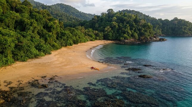 A drone captures a stunning aerial view of a secluded cove with a sandy beach and turquoise water. Lush green hills and dense tropical forest flank the coastline, creating a dramatic backdrop.