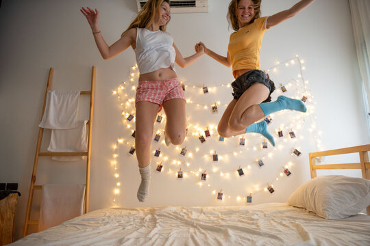 Two women joyfully jumping on a bed with string lights in the background.