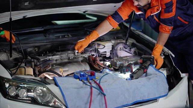 Bearded mechanic works a ratchet on the engine of a white car under a lit hood as his Asian female colleague with a green scanner leans out of frame, headlight in the foreground