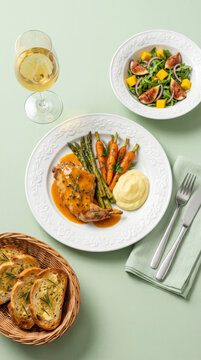 Gourmet rabbit dish in an orange sauce, served with mashed potatoes, grilled asparagus, and carrots, accompanied by a fig salad and a glass of white wine
