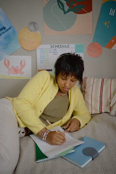 Vertical shot of African American teenage girl wearing headphones doing homework while lying down. Image suitable for online tutoring and distant learning campaigns