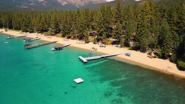 Multiple long, narrow wooden docks extend out into the water from the private properties. Footage along the picturesque sandy shore with pine tree woods of Lake Tahoe, USA.