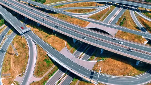 Curving ramps connect multiple levels of a highway interchange. Cars cross concrete viaducts above brown grass and narrow paths in an oblique aerial view.
