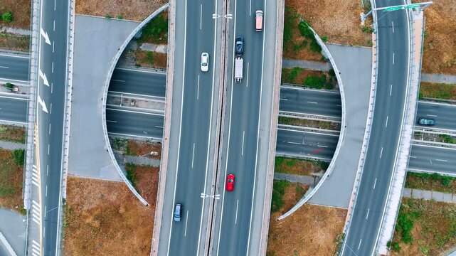 Cars travel along a highway above crossing roads. The vertical view shows white arrows and curved ramps separated by strips of brown grass.