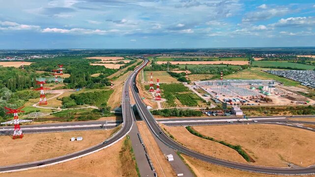 A highway crosses fields beside transmission pylons. Curved ramps fill the foreground while construction works and farmland extend toward the horizon.