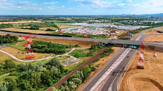 Transmission pylons flank a highway crossing. A railway follows the diagonal motorway beneath a bridge with woodland and industrial sites beyond.