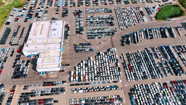 Parked vehicles surround a warehouse at a storage facility. The pale roof occupies the center while cars form dense blocks separated by marked lanes.