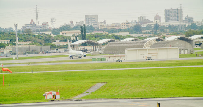 Kadena, Okinawa Prefecture, Japan - 26 April 2025: View of Kadena Air Base with a white airplane taxiing on the runway, surrounded by hangars, green grass, and urban buildings in the background.
